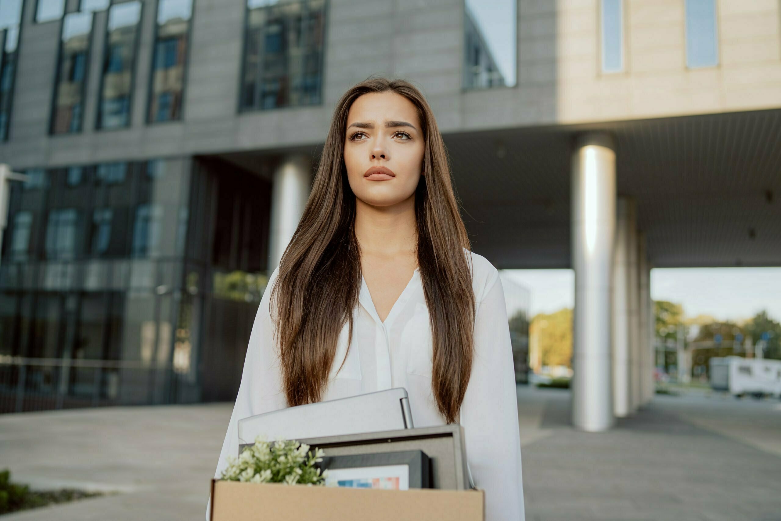 Woman stands in front of glass modern building of corporation office where she worked dismissal from position girl with sad uncertain face looks ahead holding box with packed things unemployment